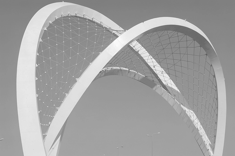 Large modern architectural arch structure with a metal mesh web design against a clear sky. Al Wahda Arches, Lusail Expressway Project, Doha, Qatar.