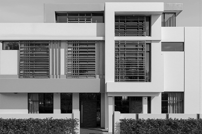 Modern two-story house facade with large rectangular windows featuring horizontal wooden slats, and a minimalist entrance surrounded by greenery. Barwa City, Qatar.